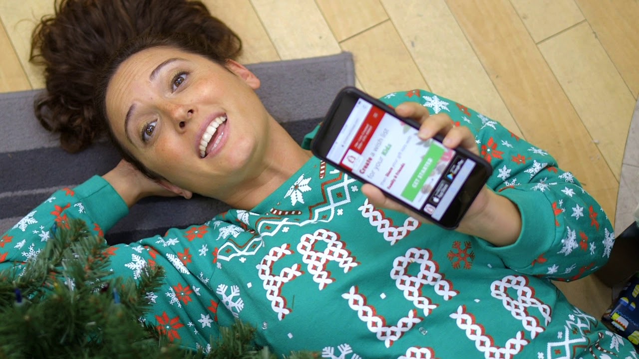 A woman in Christmas pajamas holding a phone showing ChristmasListApp